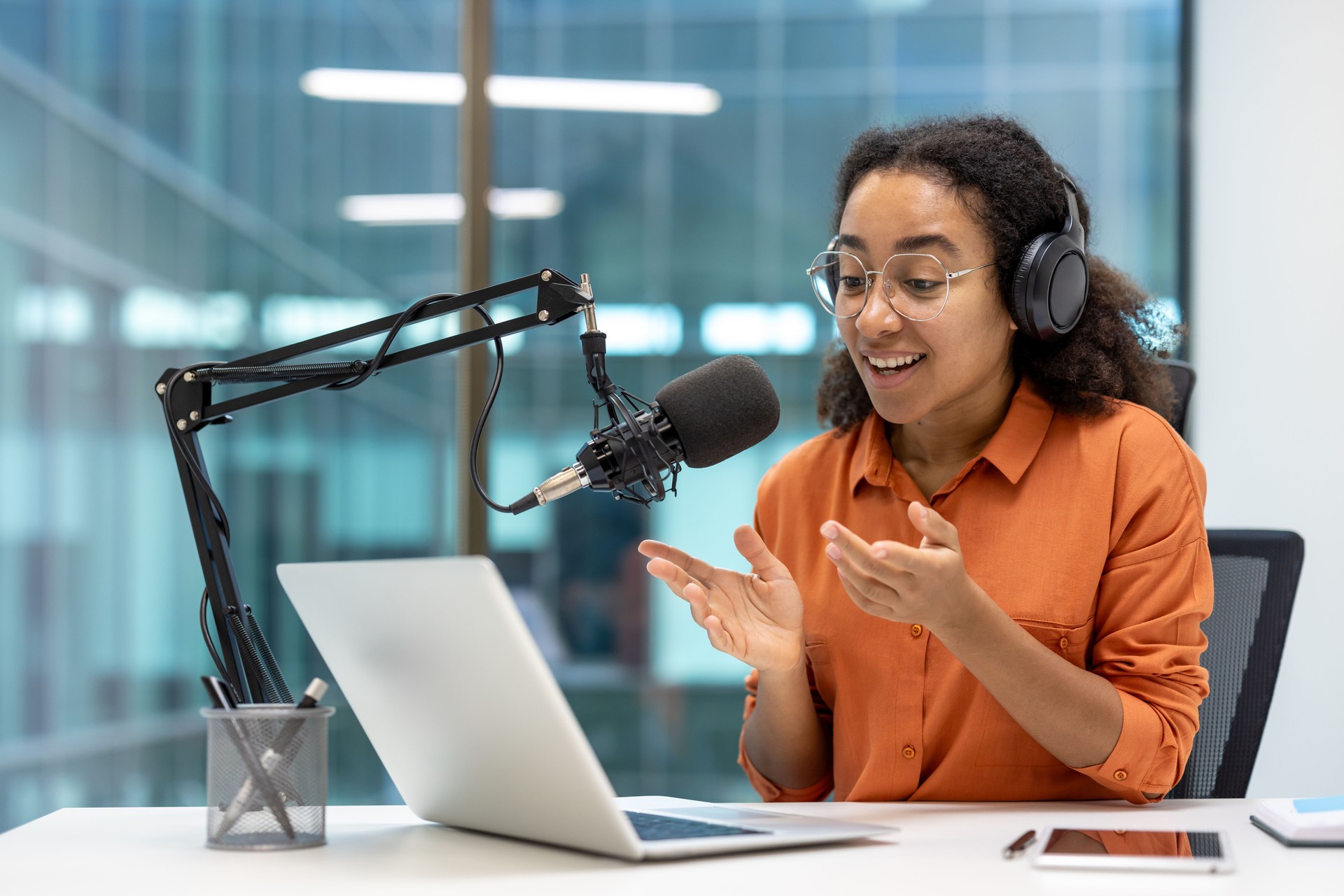 Young, cheerful woman with glasses and headphones speaks into a microphone while recording a podcast in a modern office setting.